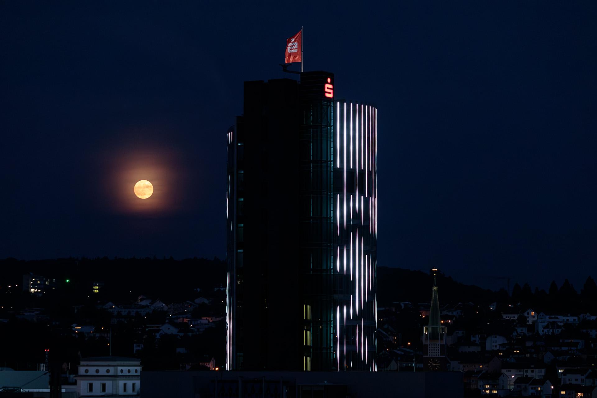 A highrise building with a red flag on top. In the background, about the lowest third is a city backdrop with mostly small buildings. To the left of the tower, the full moon shines orange through a thin layer of clouds. A highrise building with a red flag on top. In the background, about the lowest third is a city backdrop with mostly small buildings. To the left of the tower, the full moon shines orange through a thin layer of clouds.