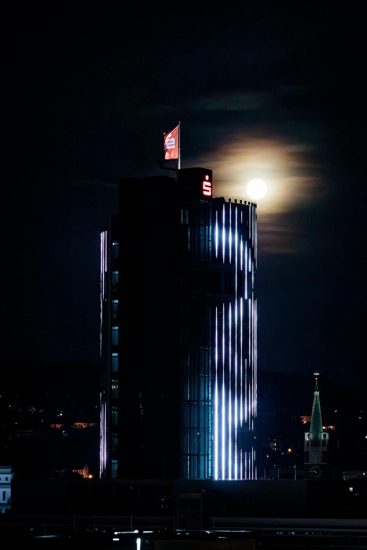 A highrise building with a red flag on top. In the background, about the lowest third is a city backdrop with mostly small buildings. Above the top edge of the tower, the full moon shines orange through a thin layer of clouds. A highrise building with a red flag on top. In the background, about the lowest third is a city backdrop with mostly small buildings. Above the top edge of the tower, the full moon shines orange through a thin layer of clouds.